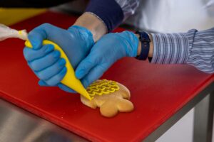 Close-up of a baker decorating a cookie with yellow icing on a red cutting board.